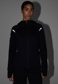 Femme portant une veste noire zippée avec des bandes réfléchissantes sur les épaules et des cordons, debout devant un fond gris avec une ombre.