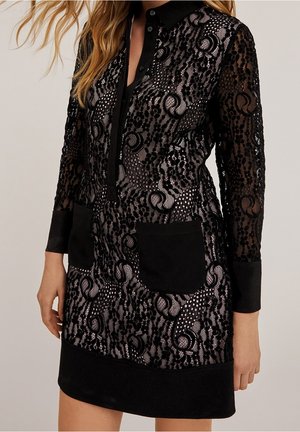 Femme portant une robe noire en dentelle à manches longues avec un col noir uni, des poignets assortis, deux poches avant et une patte de boutonnage.