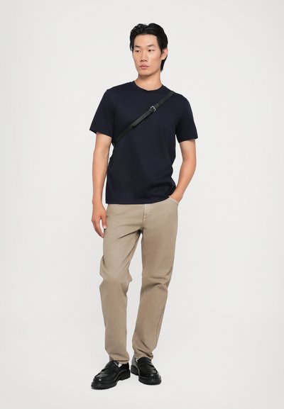T-shirt à manches courtes bleu marine foncé associé à un pantalon beige clair à coupe droite et des chaussures noires. Une sangle en bandoulière noire est portée sur la poitrine.