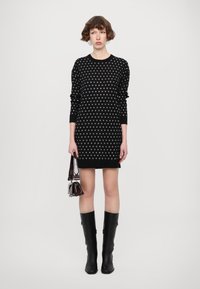 Abito nero in maglia con pois bianchi, maniche lunghe e collo rotondo. Modella che indossa stivali neri fino al ginocchio e tiene una piccola borsa.