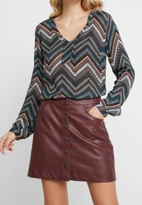 Chemise à manches longues avec un motif en zigzag vert, marron et bleu ; associée à une jupe en similicuir bordeaux avec boutons sur le devant et poches.
