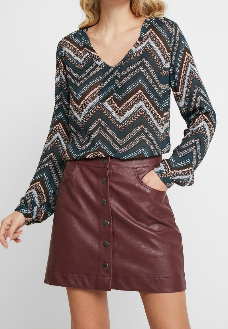 Chemise à manches longues avec un motif en zigzag vert, marron et bleu ; associée à une jupe en similicuir bordeaux avec boutons sur le devant et poches.