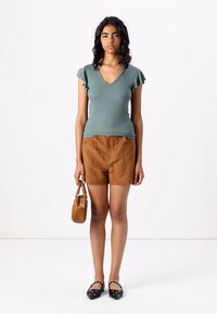 Teal ribbed top with ruffled sleeves, paired with brown suede shorts. The model holds a small orange handbag and wears black flats with accents.