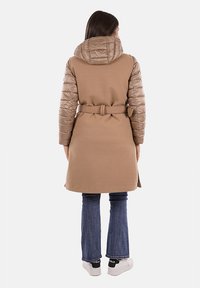Lungo cappotto beige con maniche e cappuccio marroni trapuntati, vita regolata da una cintura e una morbida texture. Indossato sopra jeans svasati scuri e sneakers bianche.