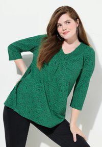 Zielony top tunikowy z dekoltem w serek i rękawami 3/4, zdobiony drobnym wzorem liści. Zestawiony z czarnymi dopasowanymi spodniami. Gładka tekstura.