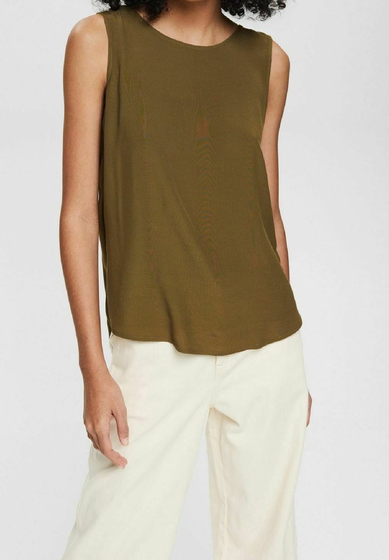 Personne portant un haut sans manches vert olive et un pantalon ample blanc cassé, debout avec une main détendue sur le côté.