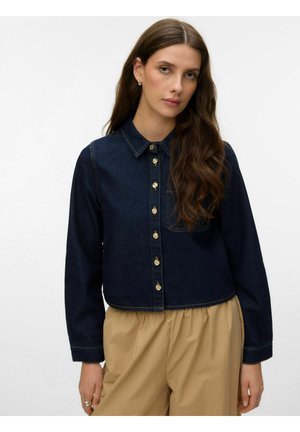 Giovane donna con lunghi capelli castani indossa una camicia di jeans blu scuro con bottoni e pantaloni beige larghi, in piedi davanti a uno sfondo chiaro e uniforme.