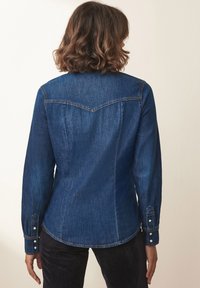 Cămașă din denim de culoare bleumarin, cu mâneci lungi, detaliu cu umeri curbati și manșete cu buton. Prezintă o siluetă ajustată și o textură subtilă.