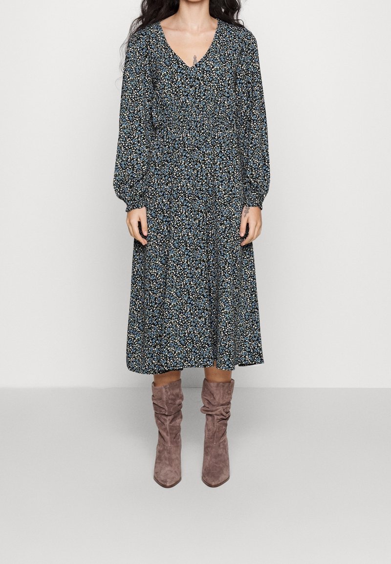 Kukallinen midi-mekko, jossa on pitkät hihat, V-pääntie, gathered-vyötärö, tummansininen pohja ja pieniä monivärisiä kukkia. Yhdistetty vaaleanruskeisiin löysiin saappaisiin.