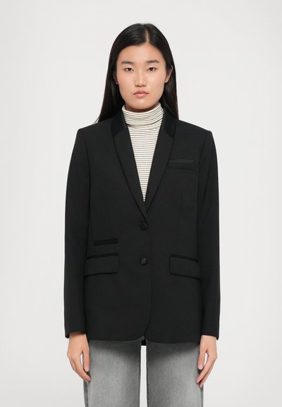 Blazer nero con rever in raso, due tasche frontali e chiusura con bottone. Indossato sopra un dolcevita a righe. Texture liscia, vestibilità strutturata.