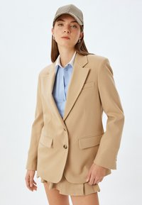 Beige skræddersyet blazer med spidse klapper, forlommer og guldknaplukning, lagd over en lyseblå skjorte. Struktur stof.