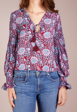 Femme portant un chemisier floral bordeaux avec des motifs bleus et roses, un lien à franges au niveau de l'encolure, et des manches cloche froncées, associé à un jean bleu.