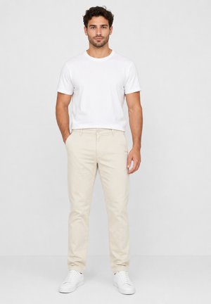 Man draagt een effen wit T-shirt, beige broek en witte sneakers, staand met één hand in de zak tegen een effen achtergrond.