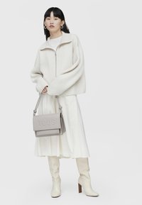 Pull zippé en tricot côtelé crème, jupe tiered blanche, bottes beiges jusqu'aux genoux et sac à bandoulière en cuir gris avec le logo "TOUS" en relief.