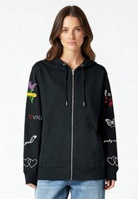 Čierna mikina na zips s prednými vreckami, s farebnými grafickými motívmi na rukávoch vrátane kvetov, sŕdc a motýľa, vyrobená z bavlny.