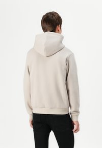 Beige Kapuzenpullover aus weichem Stoff mit einer geräumigen Kapuze und gerippten Bündchen, kombiniert mit schwarzen Jeans. Rückansicht dargestellt.