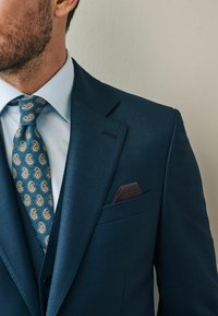 Dunkelblauer Anzug mit strukturiertem Finish, hellblaues Hemd und eine Paisleymuster-Krawatte in Teal und Gold. Enthält ein bordeauxfarbenes Einstecktuch.