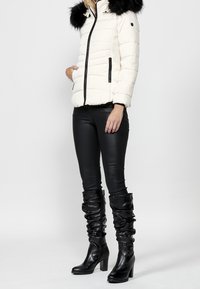 Giacca bianca imbottita con cappuccio in pelliccia sintetica nera, tasche con zip, indossata con leggins in ecopelle neri e stivali morbidi neri fino al ginocchio.