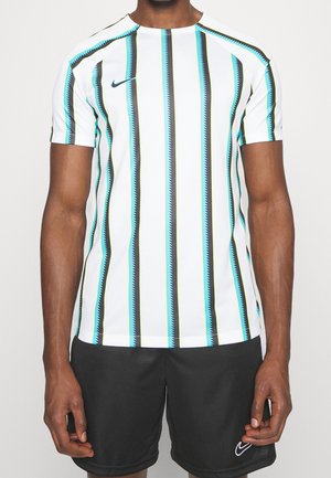 Mężczyzna w białej koszuli z krótkim rękawem z czarnymi, niebieskimi i zielonymi pionowymi zygzakowatymi paskami oraz czarnych szortach z logo Nike.