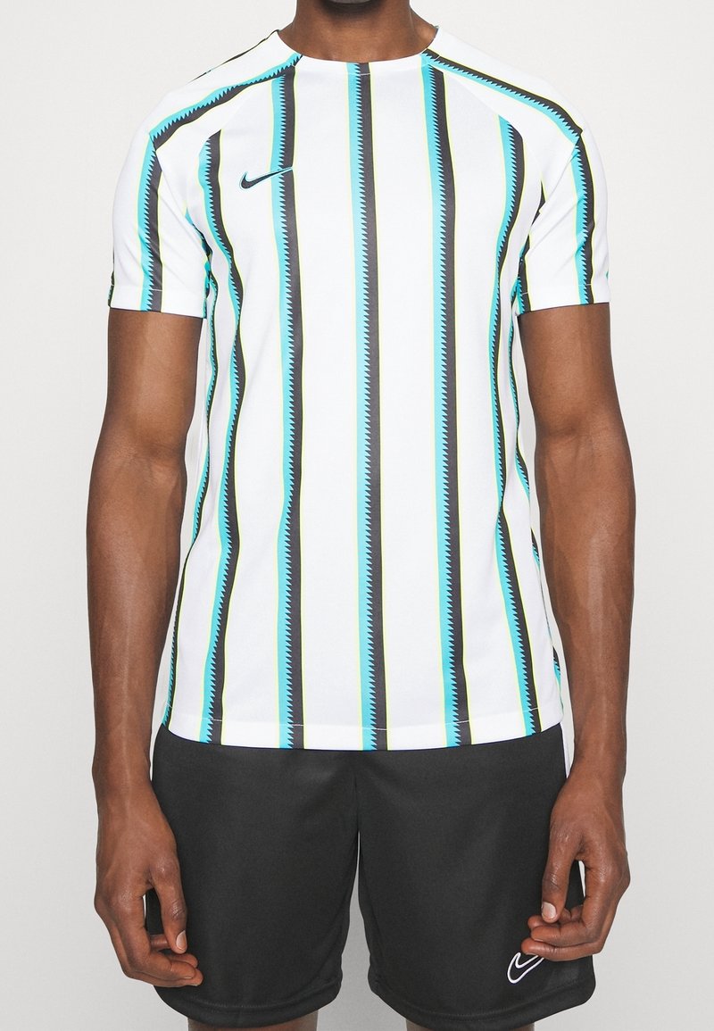 Homme portant une chemise blanche à manches courtes avec des rayures zigzag verticales noires, bleues et vertes et un short noir avec des logos Nike.