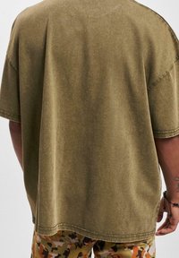 Olivově zelené oversized tričko s jemnou texturou, kulatým výstřihem a spadlými rameny, zkombinované s kalhotami s maskáčovým vzorem.