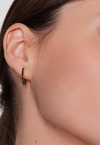 Czarne kolczyki z kamieniami szlachetnymi o zakrzywionym, hoopowym wzorze, osadzone w metalach w kolorze złota. Zbliżenie podkreśla teksturę i kształt na tle skóry.