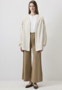 Lahki kremni kardigan z vezenimi rokavi, v kombinaciji s bež širokimi hlačami. Rjave balerinke dopolnjujejo outfit.