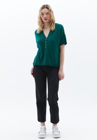 Groene kortemouwtop met een v-hals, knoopdetails en een relaxte pasvorm, gecombineerd met zwarte jeans en witte sneakers.
