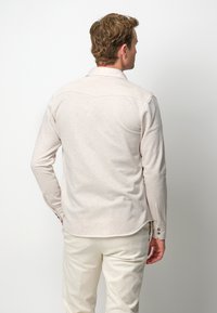 Camicia a maniche lunghe color beige chiaro con una texture morbida, colletto a punta e polsini con bottone. Presenta un orlo curvo e un dettaglio sullo yoke posteriore.