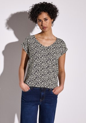 Top a maniche corte con scollatura a V di colore crema con un motivo floreale nero, abbinato a jeans blu scuro. Tessuto morbido con una vestibilità rilassata.