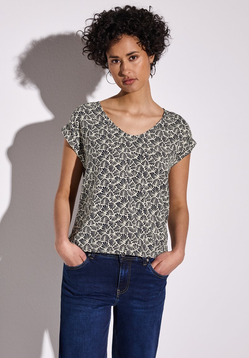 Top a maniche corte con scollatura a V di colore crema con un motivo floreale nero, abbinato a jeans blu scuro. Tessuto morbido con una vestibilità rilassata.