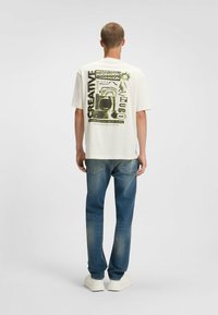 White short-sleeve t-shirt featuring bold green graphic text and imagery on the back. Paired with blue straight-leg jeans and white sneakers.