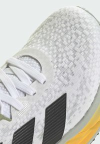 White athletic shoe featuring a textured upper, prominent black stripes, grey sole with yellow accents, and flat white laces.