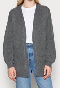 Grau gestrickte Strickjacke mit lässiger Passform, offenem Vorderteil und langen Ärmeln. Verfügt über zwei Vordertaschen und einen strukturierten Stoff. Getragen über einem weißen T-Shirt.