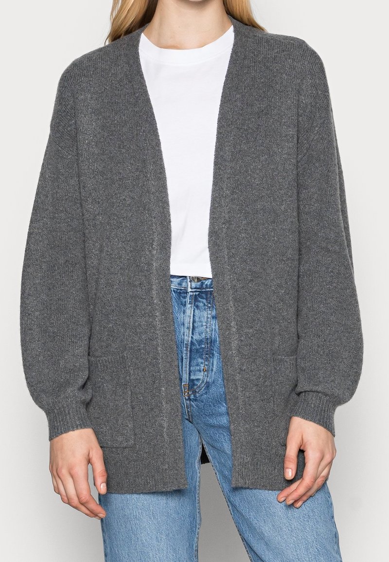 Grau gestrickte Strickjacke mit lässiger Passform, offenem Vorderteil und langen Ärmeln. Verfügt über zwei Vordertaschen und einen strukturierten Stoff. Getragen über einem weißen T-Shirt.