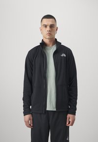 Svart zip-up jacka med hög krage, gjord av strukturerat material, med två framfickor och en liten logotyp på vänster bröst.