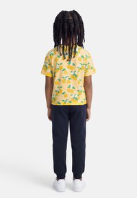 Παιδί με μακριά στριφογυριστά μαλλιά, φοράει κίτρινο πουκάμισο με σχέδιο λεμονιών, ναυτικό μπλε παντελόνι και λευκά αθλητικά παπούτσια, στέκεται με την πλάτη προς το μέρος μας σε λευκό φόντο.