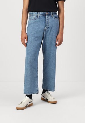 Person iført lyseblå jeans med vide ben, sorte sokker, hvide sneakers og en sort skjorte, stående mod en simpel hvid baggrund.