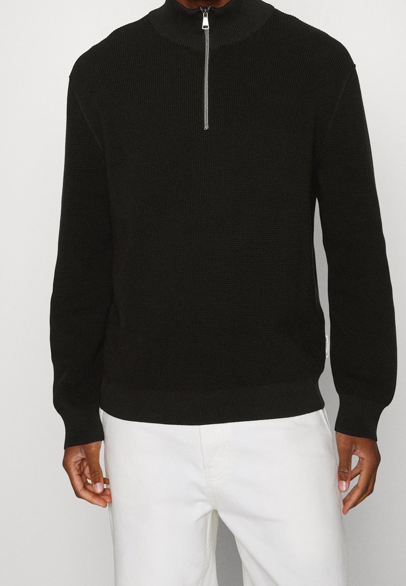 Suéter negro de media cremallera en tejido de punto texturizado, con mangas largas y un cuello ajustado. Combinado con pantalones blancos para contrarrestar.