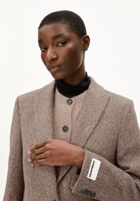 Bruine wolblend blazer met gesneden revers, textuurstof en twee knopen. Laag over een beige geribde trui met zwarte accenten.