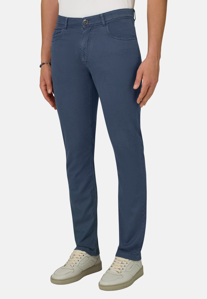 Homme portant un pantalon bleu marine coupe slim et des baskets blanc cassé, debout les mains détendues le long du corps devant un fond uni.