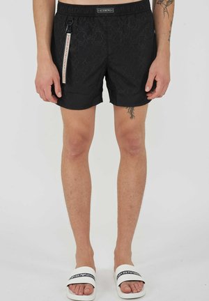 Short noir à motifs avec taille élastique et étiquette blanche pour porte-clés, porté avec des sandales blanches Emporio Armani sur une personne aux jambes nues.