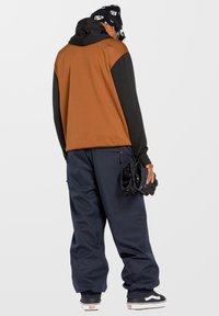 Hoodie bicolore marron et noir avec capuche à cordon de serrage, associé à un pantalon large bleu marine. Gants noirs tenus dans la main droite.