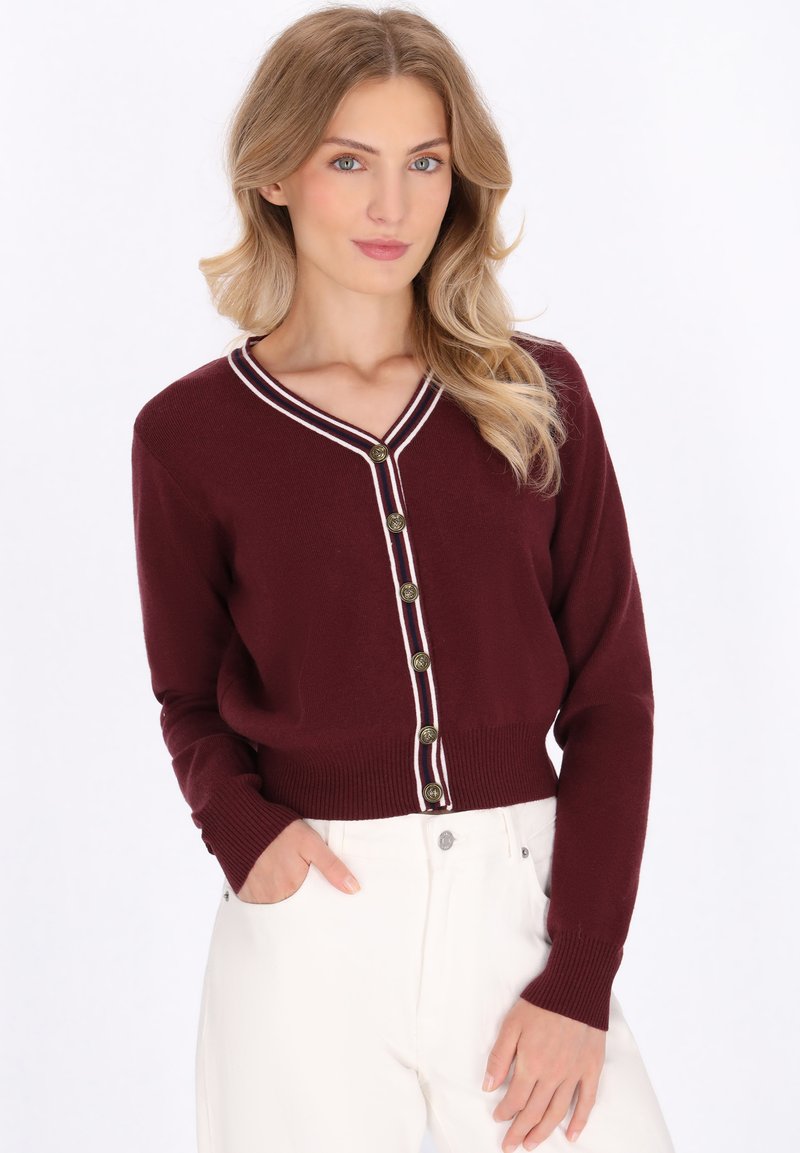 Woman with long blonde hair wearing a burgundy cardigan with gold buttons and white pants, posing with one hand in pocket.