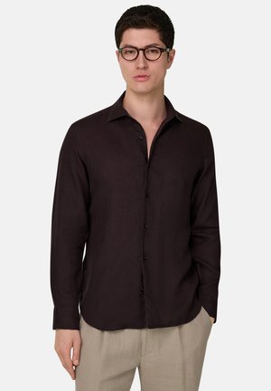 Jonge man met kort donker haar, met een zwarte ronde bril, een donkerbruine overhemd met knopen en beige broek, staand tegen een witte achtergrond.