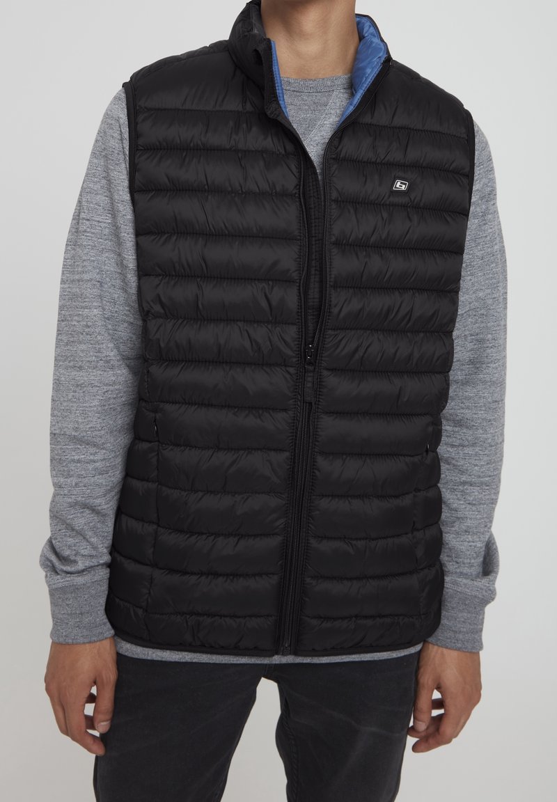 Homme portant une veste matelassée noire zippée sur une chemise grise à manches longues et un pantalon noir, debout devant un fond uni.