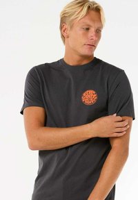 Graues kurzärmliges T-Shirt aus Baumwolle mit einem runden orangefarbenen Logo auf der linken Brust. Einfaches Design mit Rundhalsausschnitt. Glatte Textur.
