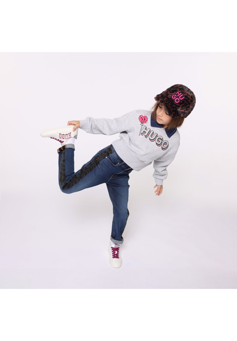 Grauer Pullover mit "HUGO"-Logo, blaue Jeans mit schwarzen Seitenstreifen, Hut mit Leopardenmuster und weiße Sneaker mit rosa Akzenten.