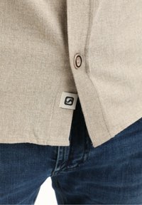 Camicia beige con tessuto testurizzato, dotata di un bottone tondo e di un piccolo etichetta con logo vicino all'orlo, abbinata a jeans blu scuri.