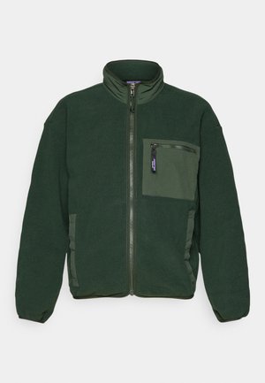 Giacca in pile verde con alto collettino, chiusura frontale con cerniera e taschino sul petto sinistro. Presenta tasche laterali e accenti in tessuto a contrasto.